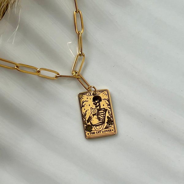Pode incluir: Colar de corrente dourada com um pingente dourado que apresenta uma ilustração em preto e branco de um esqueleto segurando um gato. O pingente tem a forma de um cartão de tarô e diz "The Cat Lover".