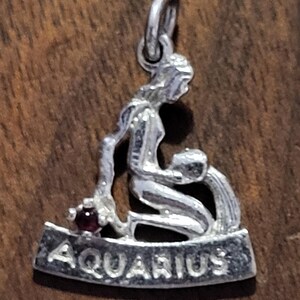 May include: Silver Aquarius charm pendant. The pendant depicts the Aquarius zodiac symbol, a woman pouring water. A small red gemstone is set near the woman. The word "AQUARIUS" is inscribed on the base of the charm.