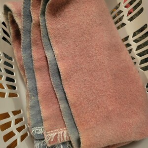 Peut inclure: Une couverture en laine rose et bleue avec une bordure frangée. La couverture est pliée en deux et se trouve dans un panier à linge blanc.