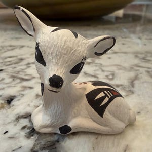 Może przedstawiać: Ceramiczna figurka jelenia w pozycji spoczynkowej. Jeleń jest głównie biały z czarnymi oznaczeniami wokół oczu, uszu i plam na ciele. Ozdobne czarno-czerwone geometryczne wzory zdobią grzbiet jelenia.