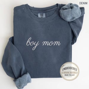 May include: A denim-coloured crewneck sweatshirt featuring the embroidered text "boy mom" in a white cursive font across the chest. The garment has a Comfort Colors label at the inner neckline and a circular badge stating "EMBROIDERED MADE TO LAST".