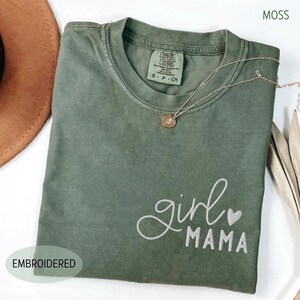 Puede incluir: Una camiseta verde musgo con el texto bordado "girl mama" en escritura blanca. La camiseta tiene cuello redondo y mangas cortas. Un collar dorado con un colgante est&aacute; drapeado sobre la camiseta. La palabra "EMBROIDERED" est&aacute; impresa abajo a la izquierda.