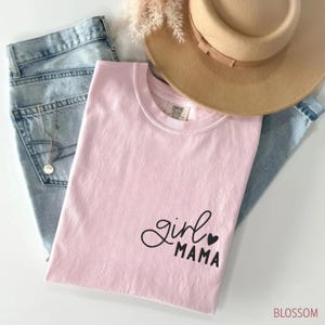 Puede incluir: Una camiseta rosa claro con las palabras "girl mama" en escritura negra y un coraz&oacute;n. La camiseta est&aacute; doblada y descansa sobre unos vaqueros azul claro y un sombrero beige. La palabra "BLOSSOM" est&aacute; en la esquina inferior derecha.