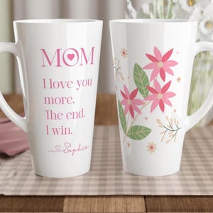 May include: Two tall white ceramic latte mugs. One mug features pink text reading MOM, I love you more. The end. I win. with the name Sophia below. The other mug displays a floral pattern with pink flowers, green leaves, and small beige sprigs.