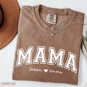 May include: A brown short-sleeved t-shirt with a crew neckline featuring the word MAMA in large white block letters. Below the text, the names Jason and Emma are printed in white script with a heart symbol between them. A silver necklace rests on top.
