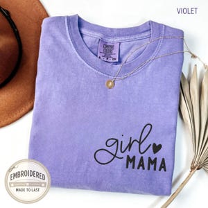 Puede incluir: Una camiseta violeta Comfort Colors con las palabras "girl MAMA" en escritura negra y un coraz&oacute;n. Un collar de oro descansa sobre la camisa. La camisa tiene un peque&ntilde;o parche bordado que dice "EMBROIDERED MADE TO LAST".