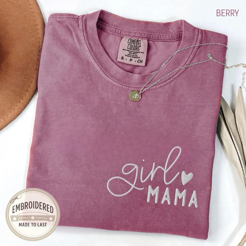 Puede incluir: Camiseta color baya con las palabras bordadas "girl MAMA" en escritura blanca y un peque&ntilde;o coraz&oacute;n. La camiseta tiene cuello redondo y mangas cortas. Un collar plateado con un colgante est&aacute; drapeado sobre la camiseta. La camiseta est&aacute; doblada sobre una superficie.