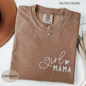 Puede incluir: Una camiseta marr&oacute;n Comfort Colors con las palabras bordadas "girl mama" en escritura blanca y un peque&ntilde;o coraz&oacute;n. Un collar plateado con un colgante descansa sobre la camiseta. La camiseta tiene cuello redondo y mangas cortas. Tambi&eacute;n se ven las palabras "MULTIPLE COLORS" y "EMBROIDERY".