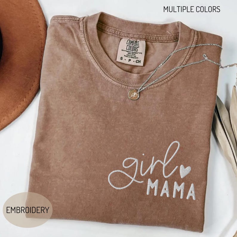 Puede incluir: Una camiseta marr&oacute;n Comfort Colors con las palabras bordadas "girl mama" en escritura blanca y un peque&ntilde;o coraz&oacute;n. Un collar plateado con un colgante descansa sobre la camiseta. La camiseta tiene cuello redondo y mangas cortas. Tambi&eacute;n se ven las palabras "MULTIPLE COLORS" y "EMBROIDERY".