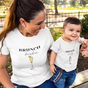 May include: A white t-shirt with the text "brunch babe" and a drawing of a mimosa glass. A white t-shirt with the text "brunch baby" and a drawing of a mimosa glass. The shirts are worn by an adult and a child.