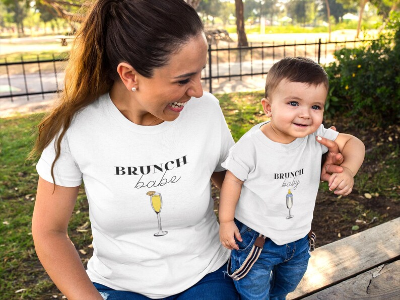 May include: A white t-shirt with the text "brunch babe" and a drawing of a mimosa glass. A white t-shirt with the text "brunch baby" and a drawing of a mimosa glass. The shirts are worn by an adult and a child.