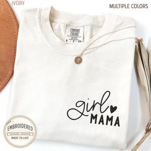 Puede incluir: Camiseta de color marfil con las palabras "girl MAMA" en escritura negra y un peque&ntilde;o coraz&oacute;n negro. Un collar dorado con un colgante redondo descansa sobre la camisa. La camiseta es de Comfort Colors.