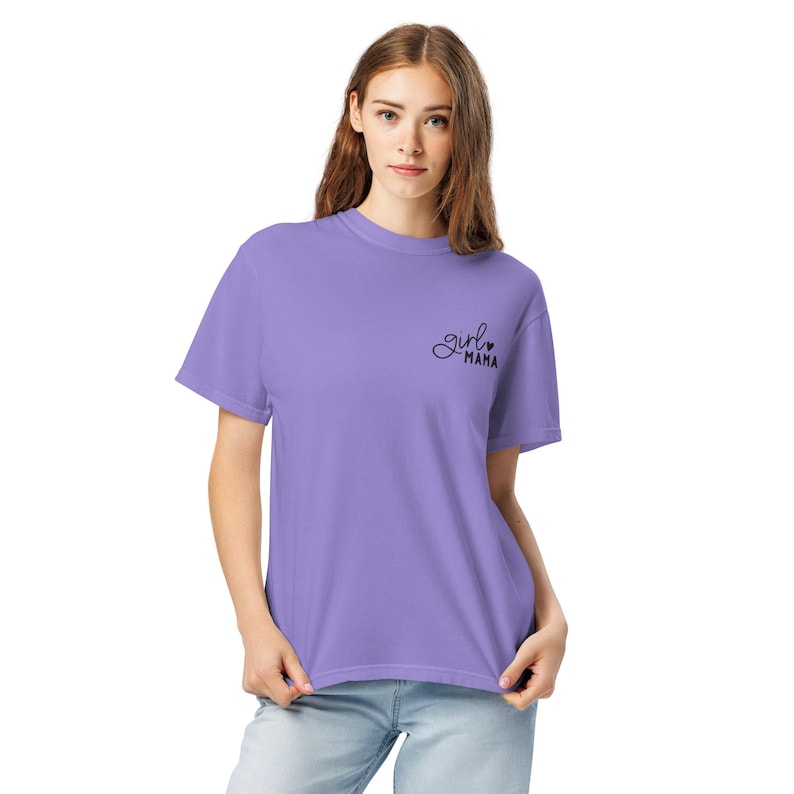 Puede incluir: Una camiseta de color lila con mangas cortas. La camiseta presenta el texto cursivo "girl mama" en la parte superior izquierda del pecho. La modelo lleva vaqueros azules. La camiseta est&aacute; hecha de un material suave y c&oacute;modo.
