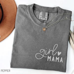 Puede incluir: Una camiseta gris con las palabras bordadas "girl MAMA" en escritura blanca y un peque&ntilde;o coraz&oacute;n. Un collar plateado con un colgante descansa sobre la camiseta. La camiseta est&aacute; doblada, con un sombrero marr&oacute;n visible en el fondo.