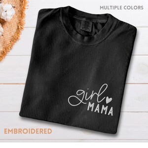 Puede incluir: Camiseta negra con texto bordado "girl mama" y un coraz&oacute;n. La camiseta se muestra sobre una superficie de madera blanca. Tambi&eacute;n son visibles las palabras "m&uacute;ltiples colores" y "bordado".