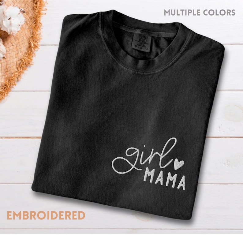Puede incluir: Camiseta negra con texto bordado "girl mama" y un coraz&oacute;n. La camiseta se muestra sobre una superficie de madera blanca. Tambi&eacute;n son visibles las palabras "m&uacute;ltiples colores" y "bordado".