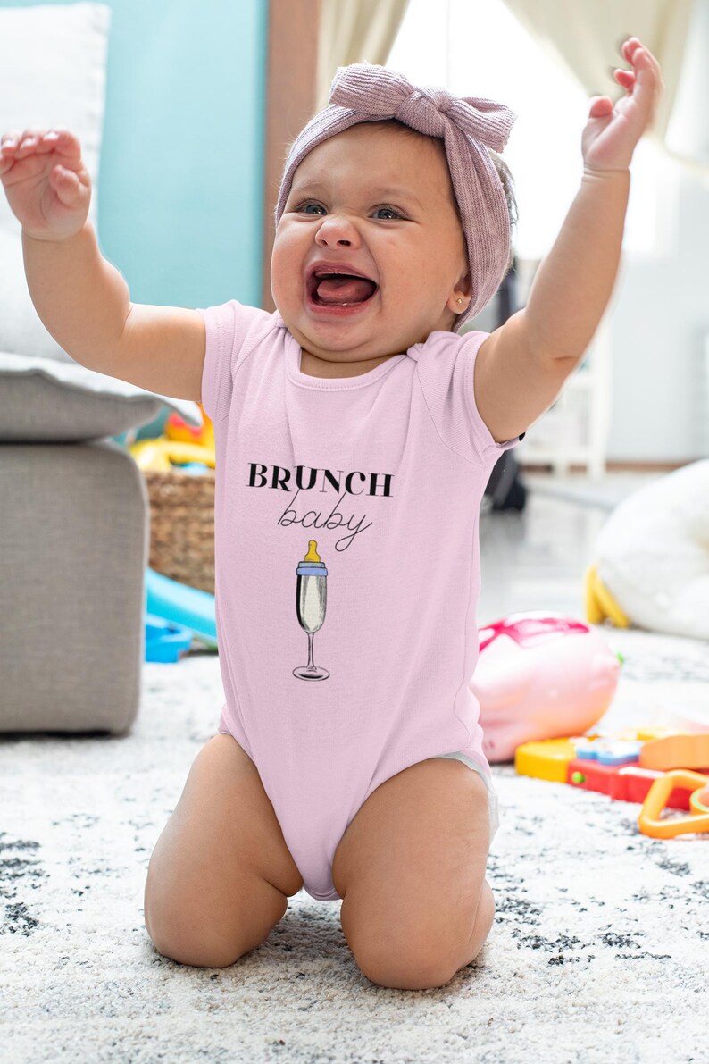May include: A pink baby onesie with the text "BRUNCH baby" and a champagne glass with a baby bottle nipple.
