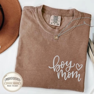 Könnte beinhalten: Ein braunes Comfort Colors T-Shirt mit den gestickten Worten "boy mom" in weißer Schrift. Eine silberne Halskette mit Anhänger liegt auf dem Shirt. Ein brauner Hut ist im Hintergrund zu sehen. Das Shirt hat ein kleines Etikett mit der Aufschrift "Comfort Colors".