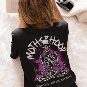 May include: Black t-shirt featuring a purple and white graphic of a skeleton sitting in a yoga pose. The text "MOTH**HOOD" is written in a circle around the skeleton. The text "DEAD TIRED, BUT STILL NAILING IT." is written below the skeleton.
