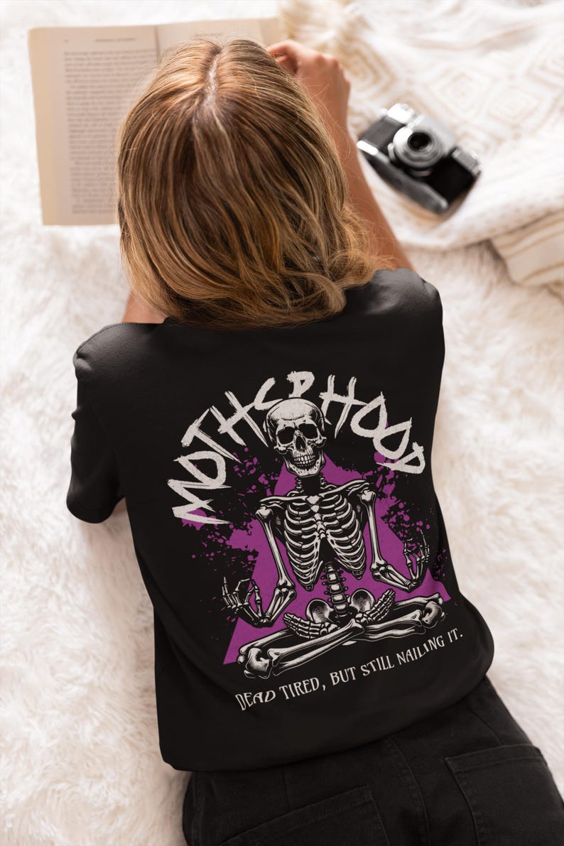 May include: Black t-shirt featuring a purple and white graphic of a skeleton sitting in a yoga pose. The text "MOTH**HOOD" is written in a circle around the skeleton. The text "DEAD TIRED, BUT STILL NAILING IT." is written below the skeleton.
