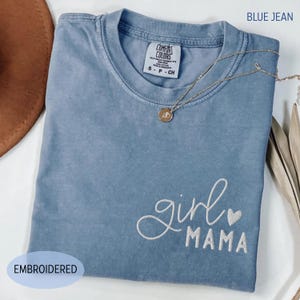 Puede incluir: Una camiseta azul vaquero con las palabras bordadas "girl mama" y un peque&ntilde;o coraz&oacute;n. La camiseta tiene cuello redondo y mangas cortas. Un collar dorado con un colgante est&aacute; drapeado sobre la camiseta. La etiqueta dice "Comfort Colors".