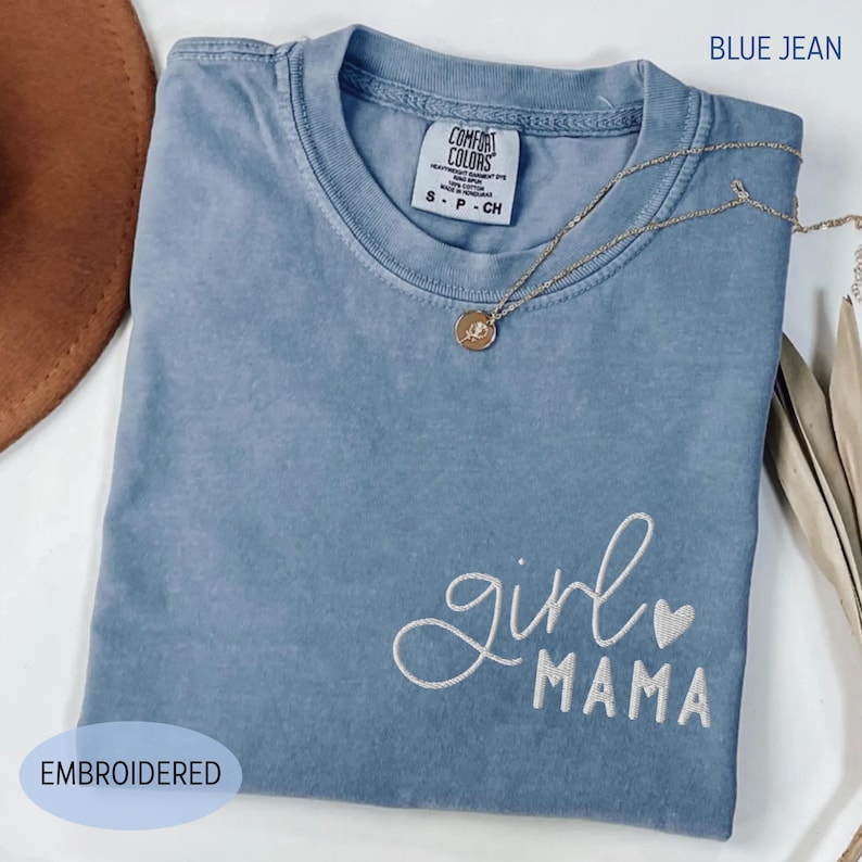 Puede incluir: Una camiseta azul vaquero con las palabras bordadas "girl mama" y un peque&ntilde;o coraz&oacute;n. La camiseta tiene cuello redondo y mangas cortas. Un collar dorado con un colgante est&aacute; drapeado sobre la camiseta. La etiqueta dice "Comfort Colors".