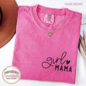 Puede incluir: Una camiseta rosa con las palabras "girl MAMA" en escritura negra y un coraz&oacute;n. La camiseta tiene cuello redondo y mangas cortas. Un collar dorado con un colgante est&aacute; drapeado sobre la camiseta. La palabra "CRUNCHBERRY" est&aacute; impresa en la parte superior.