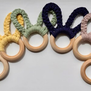 May include: A collection of seven macrame bunny ear baby teethers in various colors, including brown, yellow, green, navy, pink, and white. Each teether features a wooden ring and a knotted macrame design, perfect for soothing teething babies.