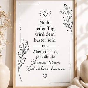 May include: A framed wall art print featuring black text on a white background. The text reads: Nicht jeder Tag wird dein bester sein. Aber jeder Tag gibt dir die Chance, deinem Ziel näherzukommen. It includes black line art of leaves and hearts.