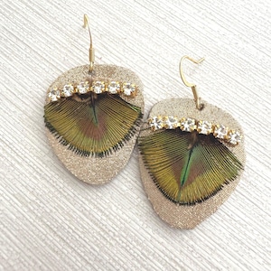 Może przedstawiać: Para ręcznie robionych kolczyków w kształcie łzy. Kolczyki mają złotą bazę z tkaniny, ozdobioną zielono-brązowym wzorem piór. Rząd przezroczystych kryształków dodaje blasku. Złote haczyki do uszu dopełniają projekt.