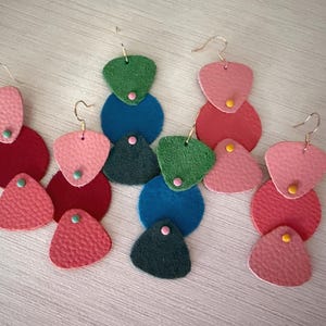 May include: Four pairs of layered drop earrings made of textured leather pieces in various shapes including circles and triangles. The colors include shades of pink, red, green, and blue, accented with small metal studs and gold-toned ear wires.