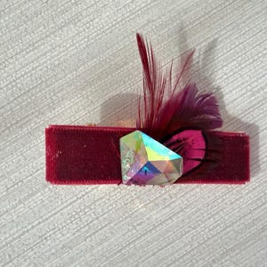 May include: A rectangular hair clip covered in deep burgundy velvet fabric. The accessory is adorned with a faceted, iridescent crystal-like gem and a cluster of dark burgundy and purple feathers extending from the side of the central stone.