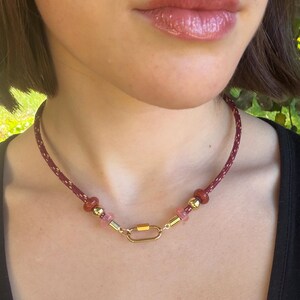 Op de afbeelding: Een ketting met een bordeauxrood koord met goudkleurige spikkels, geaccentueerd door twee rode kralen, twee goudkleurige bollen, twee doorschijnende roze kralen en twee goudkleurige cilinders, in het midden voorzien van een ovale karabijnsluiting.