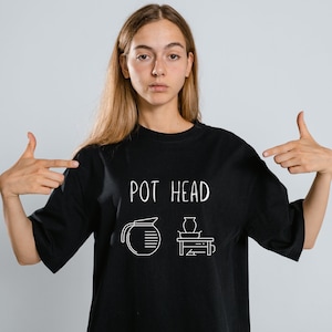 May include: A person wearing a black t-shirt with the text "POT HEAD" and a graphic of a pitcher and a vase. They are pointing at the shirt with both hands.