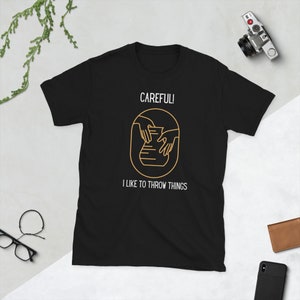 May include: Black t-shirt with a gold outline of hands shaping clay on a pottery wheel. The text "CAREFULL! I LIKE TO THROW THINGS" is printed below the image.