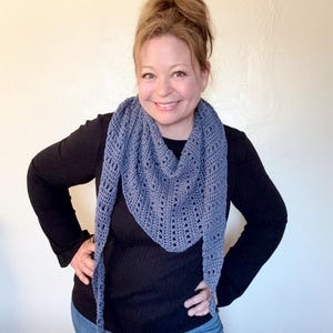 May include: A periwinkle blue crocheted shawl with a triangular shape and openwork design. The shawl is worn around the neck and drapes down the front. The shawl is made of a soft, lightweight yarn.