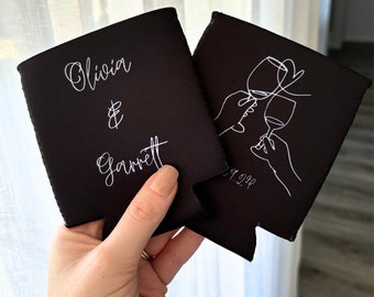 Puede incluir: Dos portavasos negros con texto blanco. Un portavasos dice "Olivia & Garrett" y el otro tiene un dibujo lineal de dos manos sosteniendo copas de vino y la fecha "09.09.22".