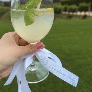 May include: A stemmed glass filled with a light yellow beverage, garnished with mint and a lemon slice. A white ribbon with the text "RBC Christmas Party 2024" is tied to the base of the glass. The background features a green lawn and a vineyard.
