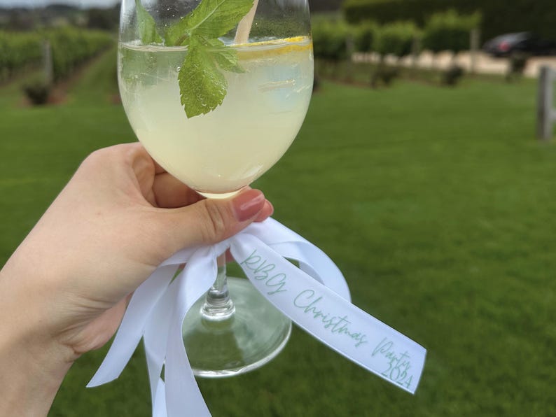 May include: A stemmed glass filled with a light yellow beverage, garnished with mint and a lemon slice. A white ribbon with the text "RBC Christmas Party 2024" is tied to the base of the glass. The background features a green lawn and a vineyard.
