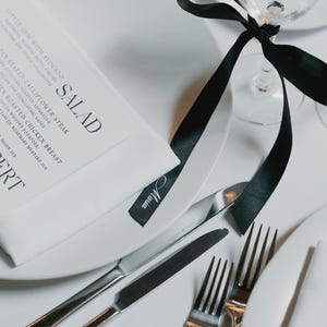 May include: Elegant table setting with white plates, silverware, and a menu. A black ribbon is tied around a wine glass. The menu lists salad, main course, and dessert options. A small tag with the name "Maria" is visible.
