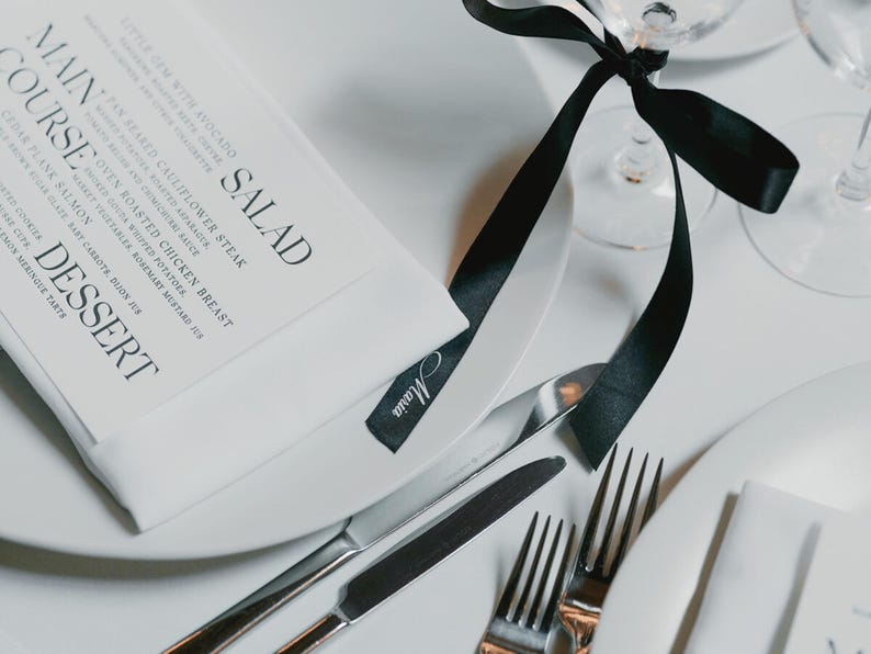 May include: Elegant table setting with white plates, silverware, and a menu. A black ribbon is tied around a wine glass. The menu lists salad, main course, and dessert options. A small tag with the name "Maria" is visible.