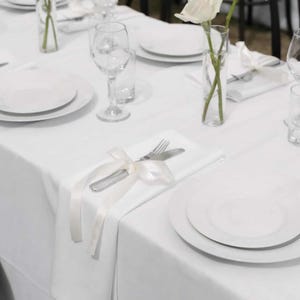 May include: A formal table setting with white plates, silverware, and wine glasses. A white tablecloth covers the table, and a white ribbon adorns the silverware. A single white rose in a clear vase adds a touch of elegance.