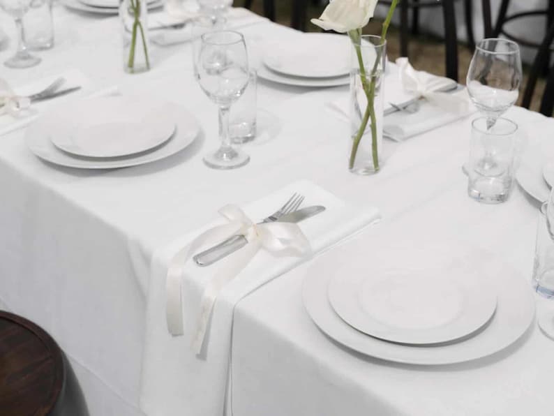 May include: A formal table setting with white plates, silverware, and wine glasses. A white tablecloth covers the table, and a white ribbon adorns the silverware. A single white rose in a clear vase adds a touch of elegance.