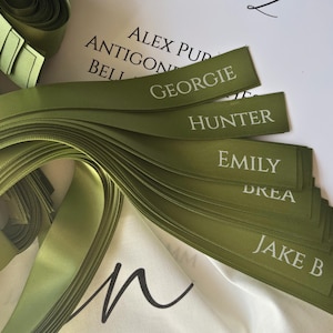 以下が含まれることがあります： 白文字で「Georgie」、「Hunter」、「Emily」などの名前が書かれた、オリーブグリーンのサテンリボン。リボンはきちんと配置され、背景にはリボンのロールが見えます。名前はクラシックなフォントで印刷されています。