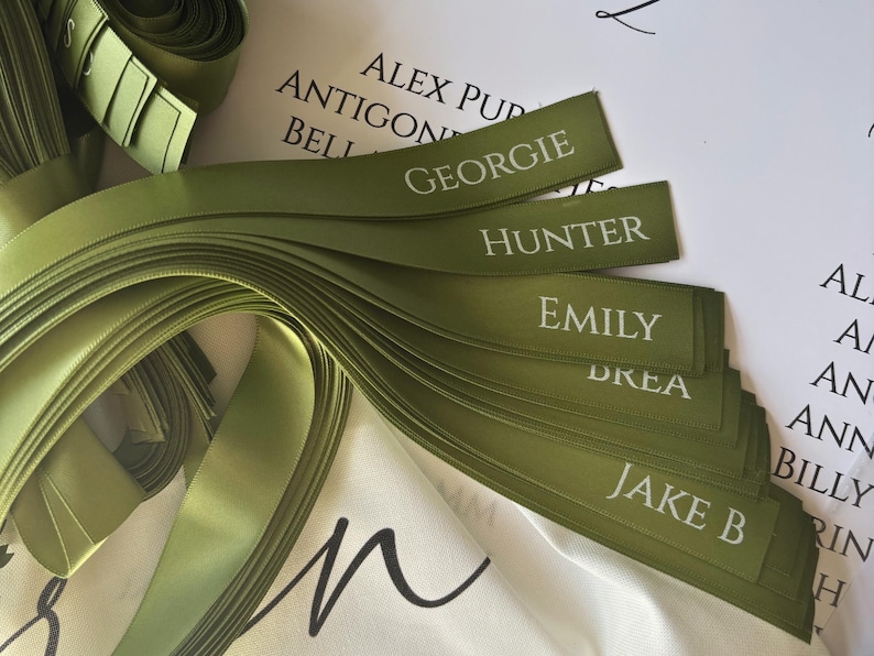 May include: Olive green satin ribbons with white text, featuring names like "Georgie," "Hunter," and "Emily." The ribbons are neatly arranged, with a roll of ribbon visible in the background. The names are printed in a classic font.
