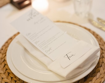 Könnte beinhalten: Ein weißes Gedeck mit einem geflochtenen Platzdeckchen, einem weißen Teller, einer weißen Serviette und einer Speisekarte mit dem Wort "Tom" darauf.