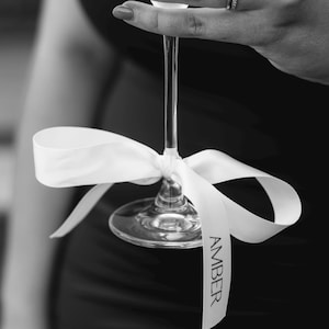 May include: A clear champagne flute with a white ribbon tied around the stem. The ribbon has the name "AMBER" printed on it in black. The image is in black and white.