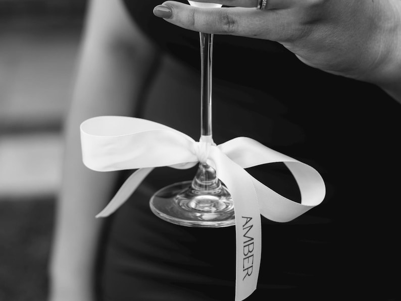 May include: A clear champagne flute with a white ribbon tied around the stem. The ribbon has the name "AMBER" printed on it in black. The image is in black and white.