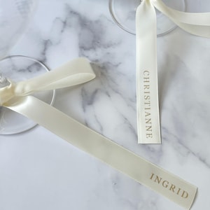 May include: Ivory satin ribbons with gold text reading "CHRISTIANNE" and "INGRID" are tied to the stems of clear wine glasses. The ribbons are arranged on a white and gray marble surface, suggesting a wedding or event.
