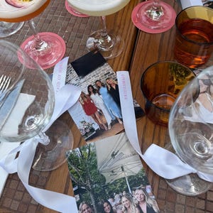 May include: A table setting with cocktails, glassware, and photos. Clear wine glasses with white ribbons, a fork and knife, and amber-colored glasses are visible. Pink coasters and photos with the text "Maid of Honor" are also present.